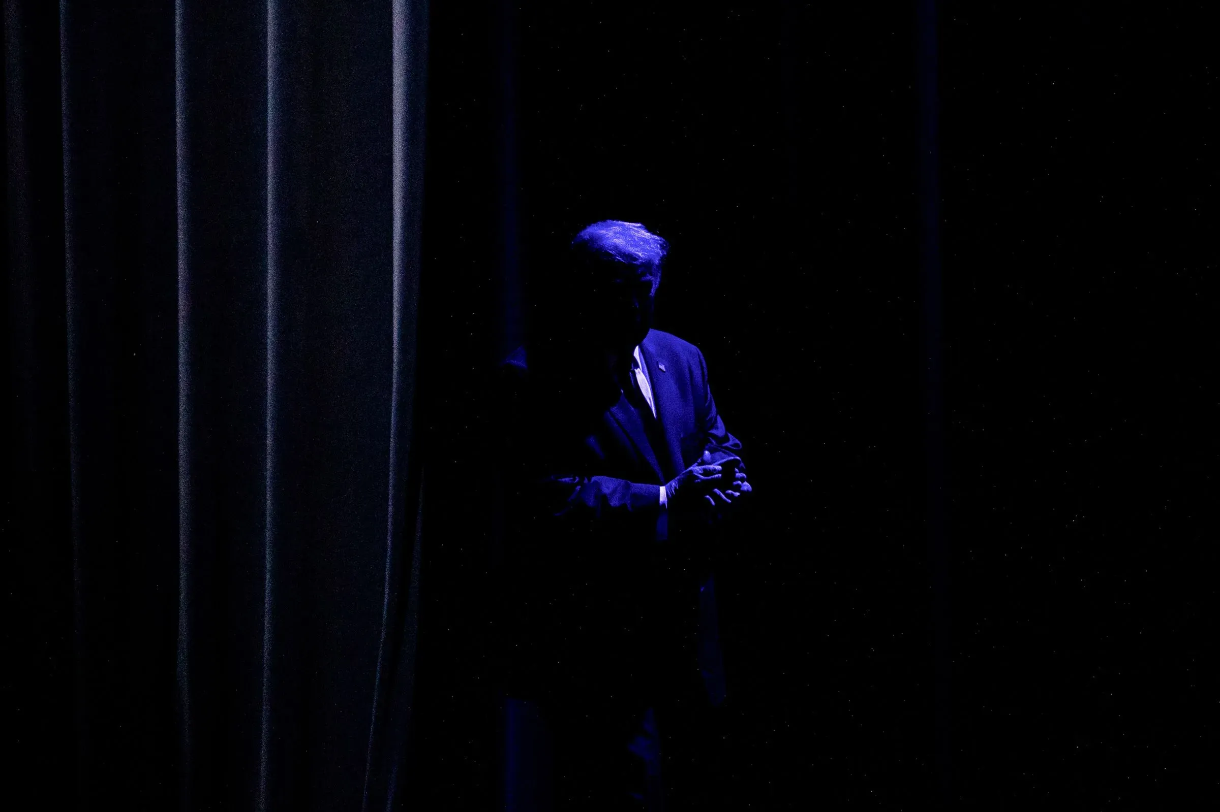 شخصية سياسية بارزة تقف في الظل خلف ستارة، مما يعكس التوترات السياسية الحالية وتأثيرها على المجتمع الأمريكي.