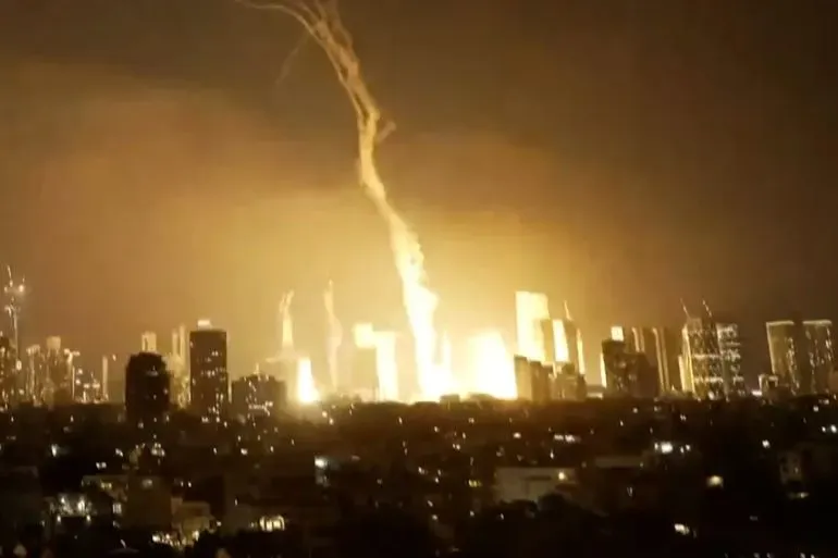 تظهر الصورة انفجارًا ضخمًا يضيء سماء مدينة في الليل، مما يعكس تصاعد التوترات في الصراع الإسرائيلي-الإيراني.