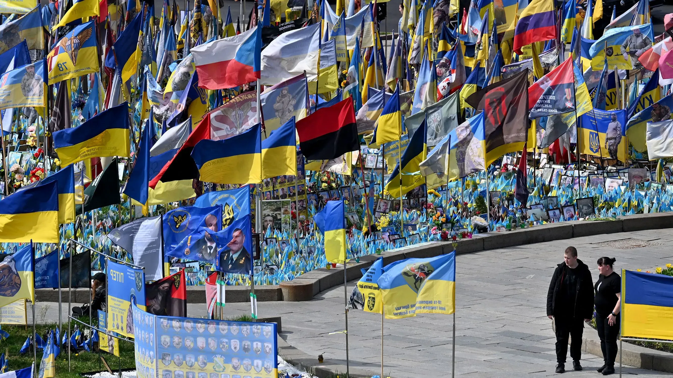 تظهر الصورة مجموعة من الأعلام الأوكرانية والرايات الأخرى في موقع تذكاري، حيث يسير شخصان بالقرب من الأعلام في مشهد يعكس التضامن خلال النزاع.