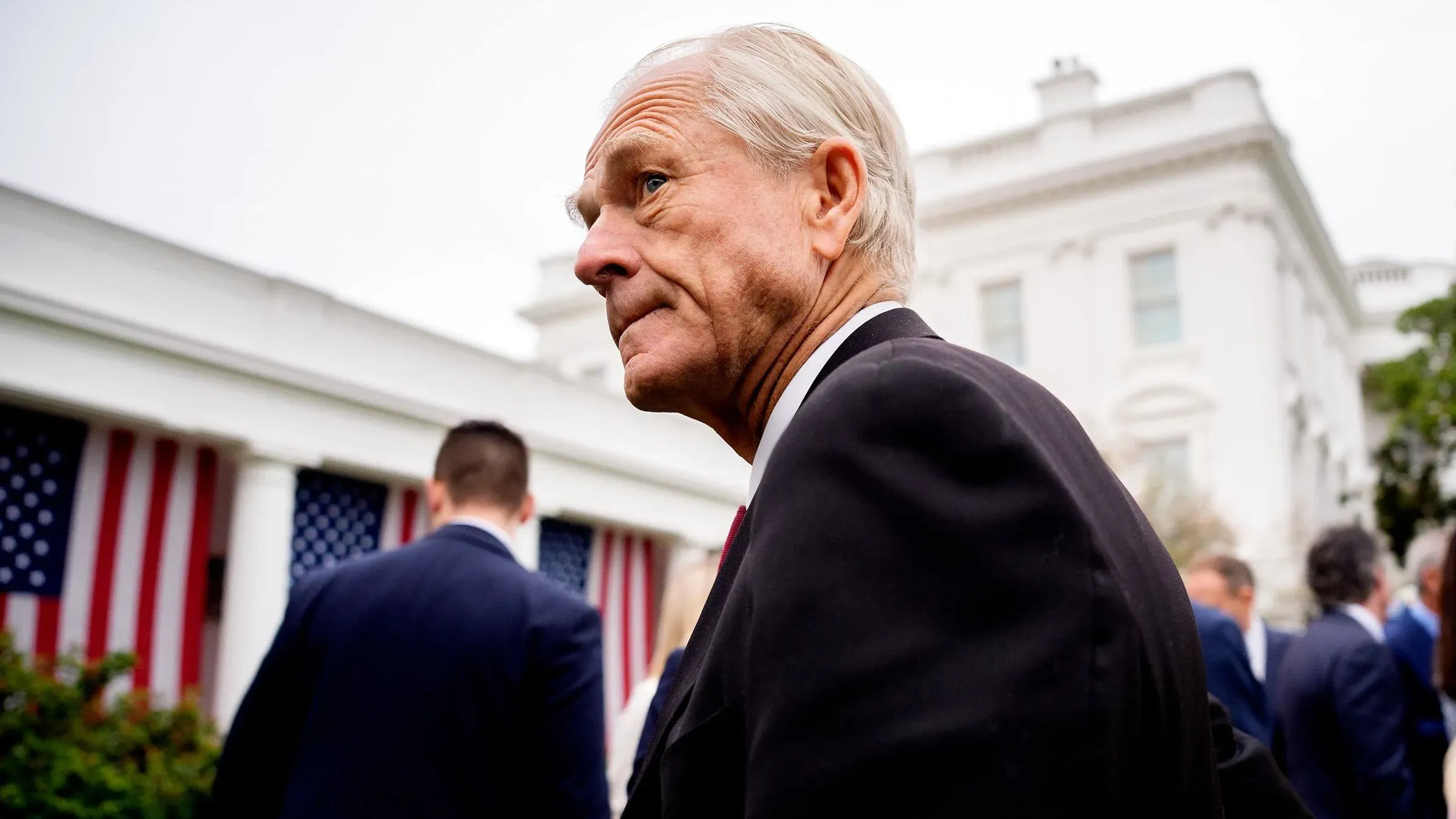 بيتر نافارو، مستشار التجارة في البيت الأبيض، يسير بجانب مبنى حكومي مع أعلام أمريكية، معبرًا عن تأثيره المستمر في السياسة التجارية.