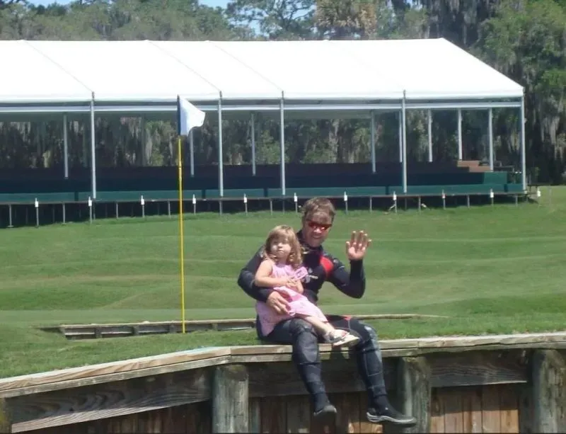 جيم بيست يرتدي بدلة غوص ويجلس مع فتاة صغيرة على حافة ملعب جولف TPC Sawgrass، مع العلم الأبيض في الخلفية.