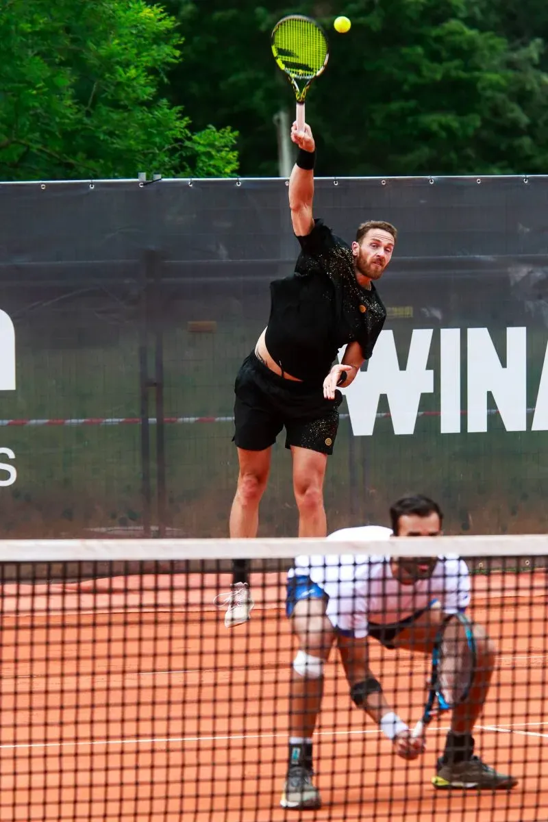 لاعب تنس يقوم بإرسال الكرة خلال مباراة على أرضية ترابية، مع التركيز على تقنياته في الإرسال السريع.