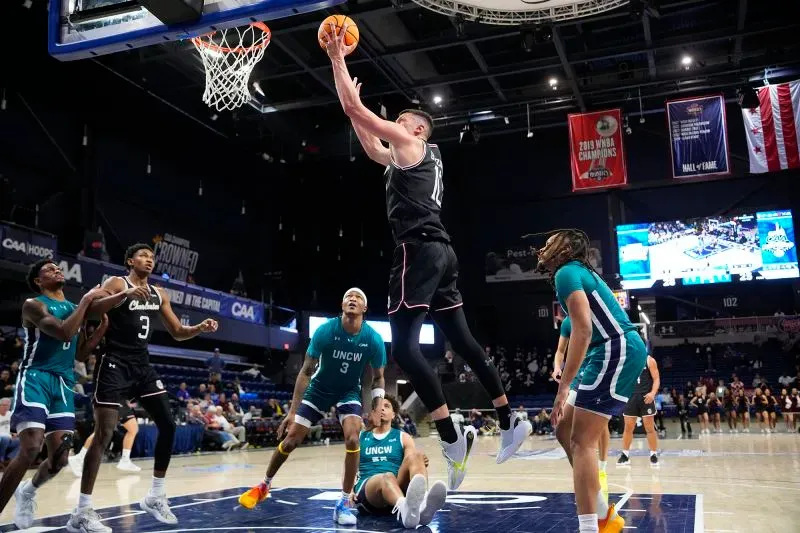 لاعب من فريق توسون يقوم بقفزة ساحقة نحو السلة خلال مباراة بطولة الرابطة الرياضية الساحلية (CAA) لكرة السلة، مع وجود لاعبين من جامعة نورث كارولينا ويلمنجتون في الخلفية.