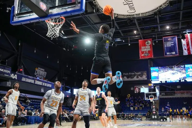 لاعب كرة سلة يقفز عالياً لتسجيل سلة خلال مباراة في بطولة CAA، مع تواجد لاعبين آخرين في الخلفية وجمهور متحمس.