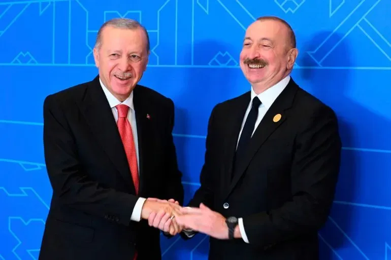 لقاء بين الرئيس التركي رجب طيب أردوغان ورئيس أذربيجان إلهام علييف، حيث يتصافحان مبتسمين، مع خلفية زرقاء.