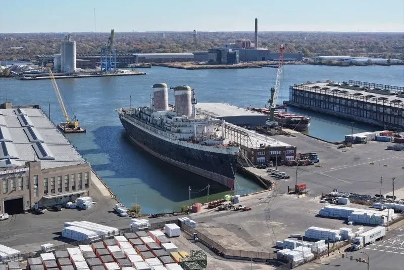 باخرة SS United States موقوفة في رصيف فيلادلفيا، محاطة بمرافق الميناء والمباني، في انتظار الإبحار نحو خليج المكسيك لتصبح شعابًا مرجانية.