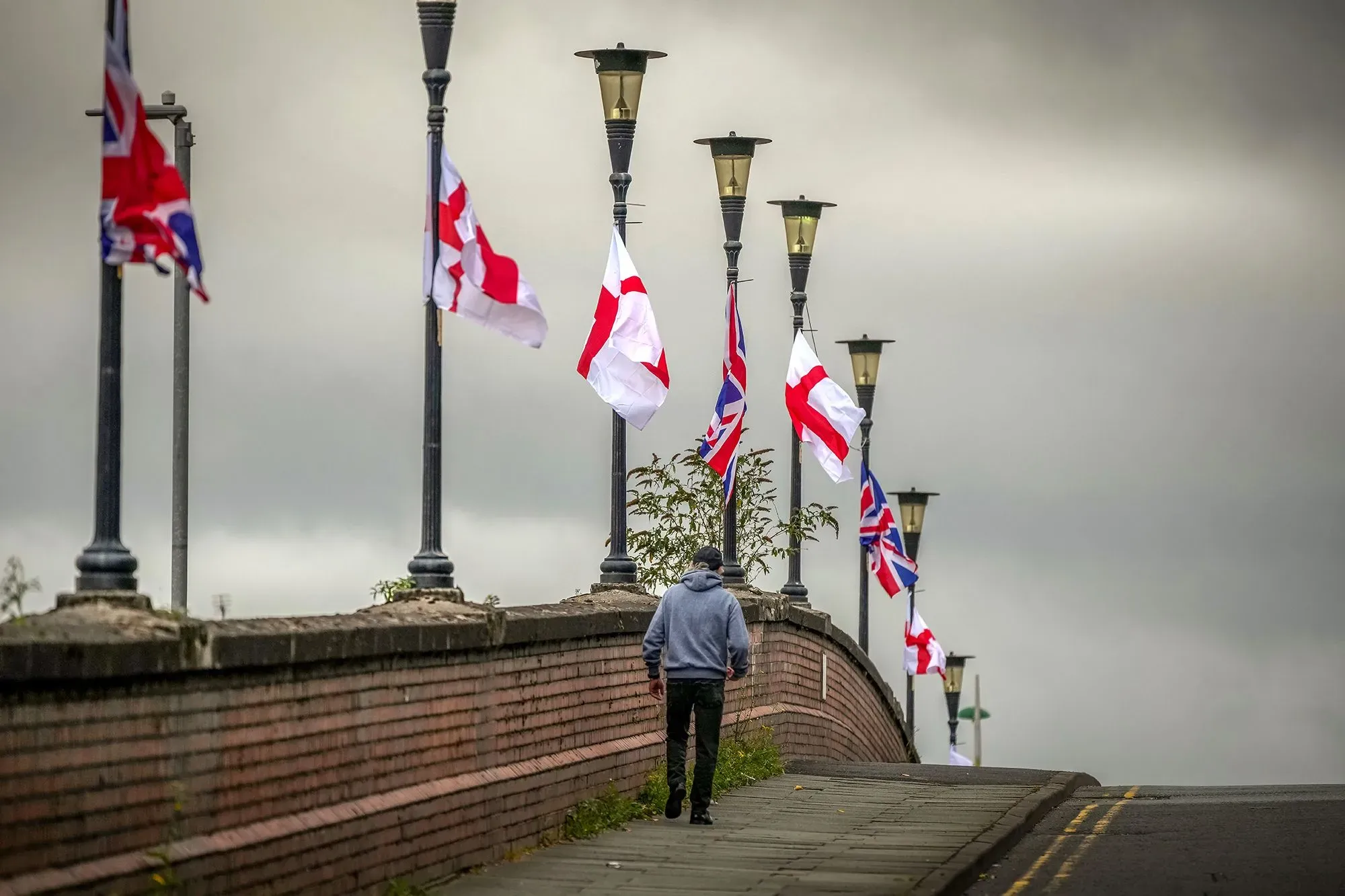 أعلام المملكة المتحدة وعلم إنجلترا ترفرف على أعمدة الإنارة في مدينة إنجليزية، مع شخص يمشي على الرصيف، مما يعكس التوترات الوطنية الحالية.