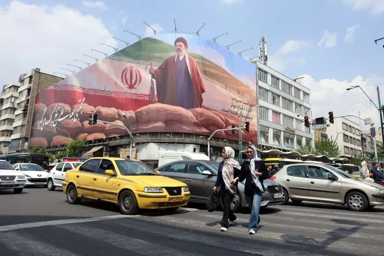 صورة لملصق ضخم يظهر المرشد الإيراني الجديد آية الله مجتبى خامنئي، مع العلم الإيراني، في وسط طهران، بينما تعبر امرأتان الشارع.