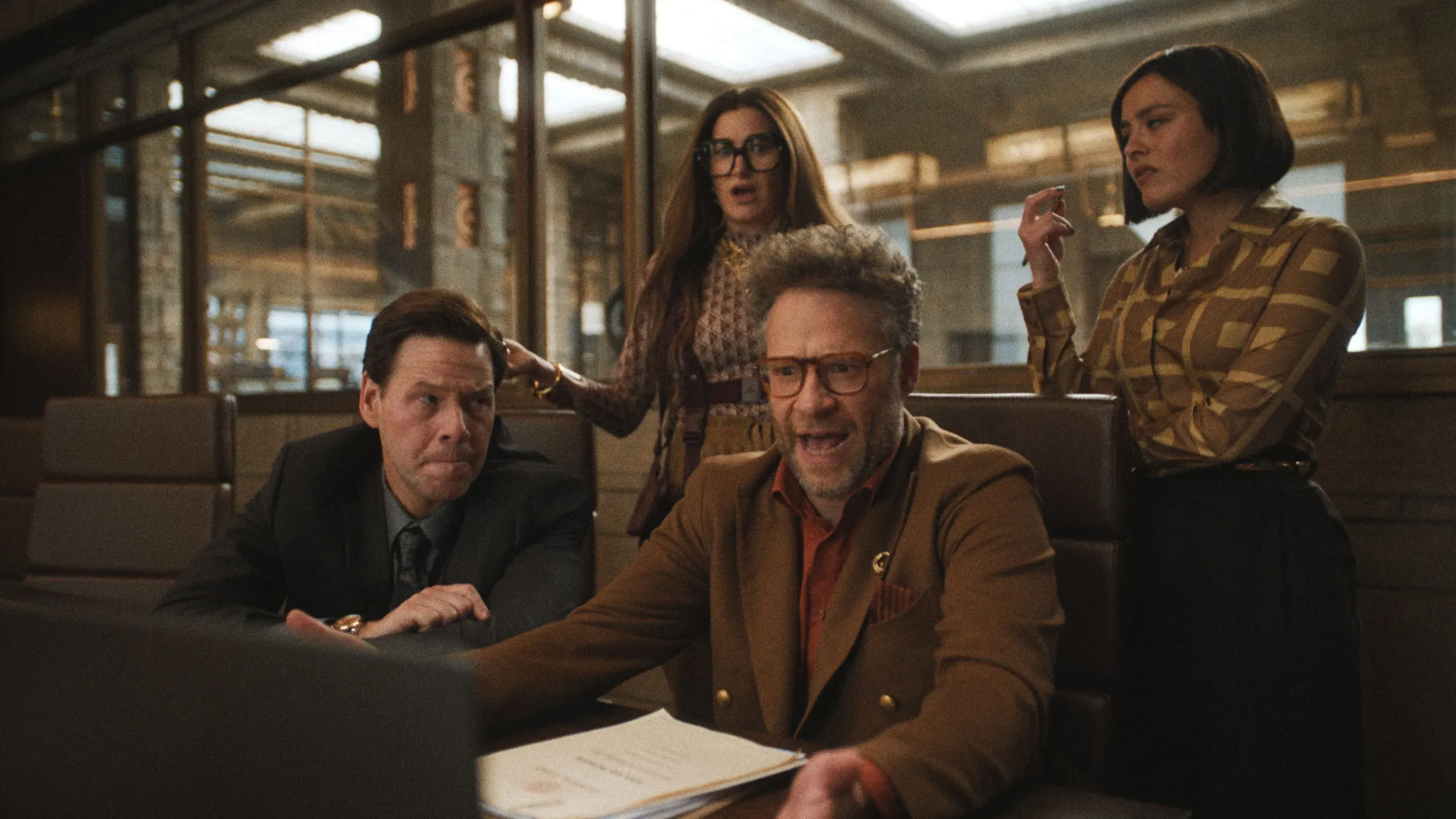 مجموعة من الشخصيات في اجتماع مكتبي، مع تعبيرات متوترة، تتفاعل مع عرض تقديمي على الكمبيوتر في استوديو أفلام.