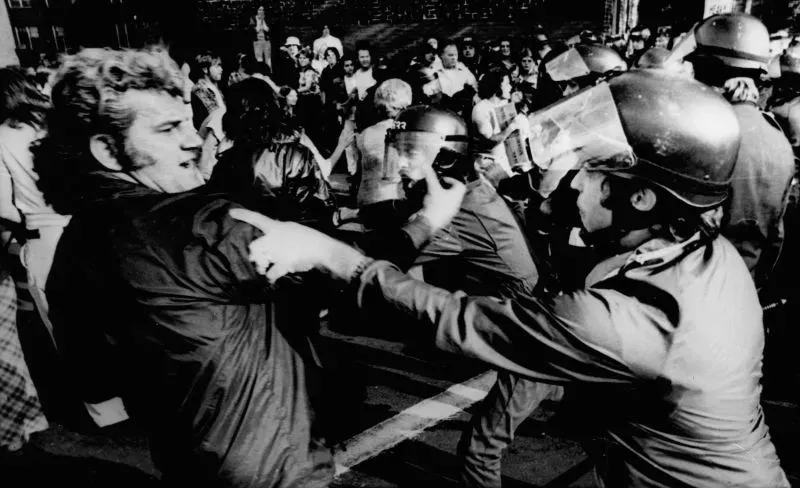 مشهد من احتجاجات في السبعينات، يظهر صدامًا بين متظاهرين ورجال شرطة، مما يعكس التوترات الاجتماعية حول العدالة العرقية.