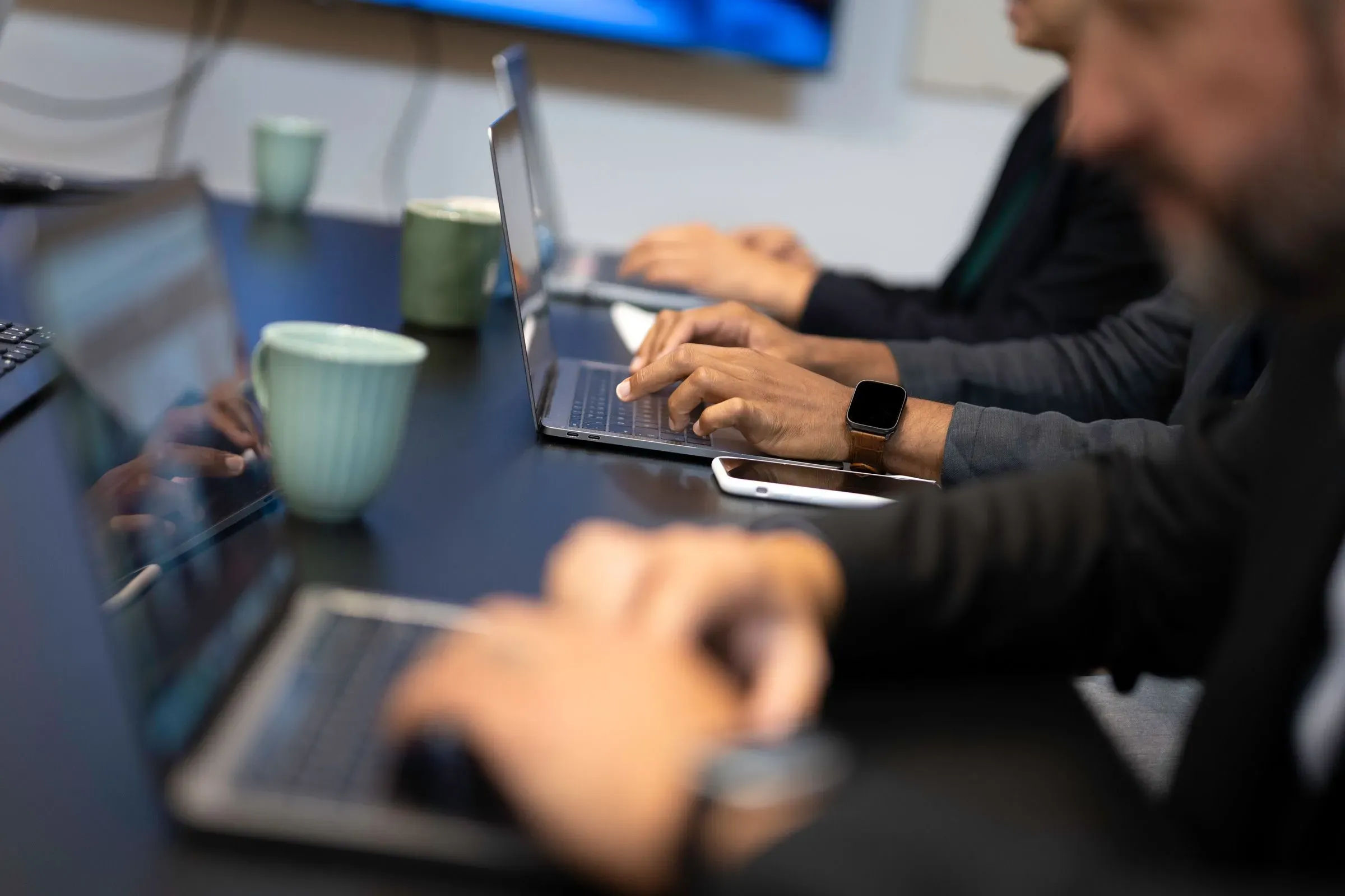 أشخاص يعملون على أجهزة الكمبيوتر المحمولة في بيئة مكتبية، مع أكواب شاي أو قهوة، يعكسون تأثير الذكاء الاصطناعي على سوق العمل.