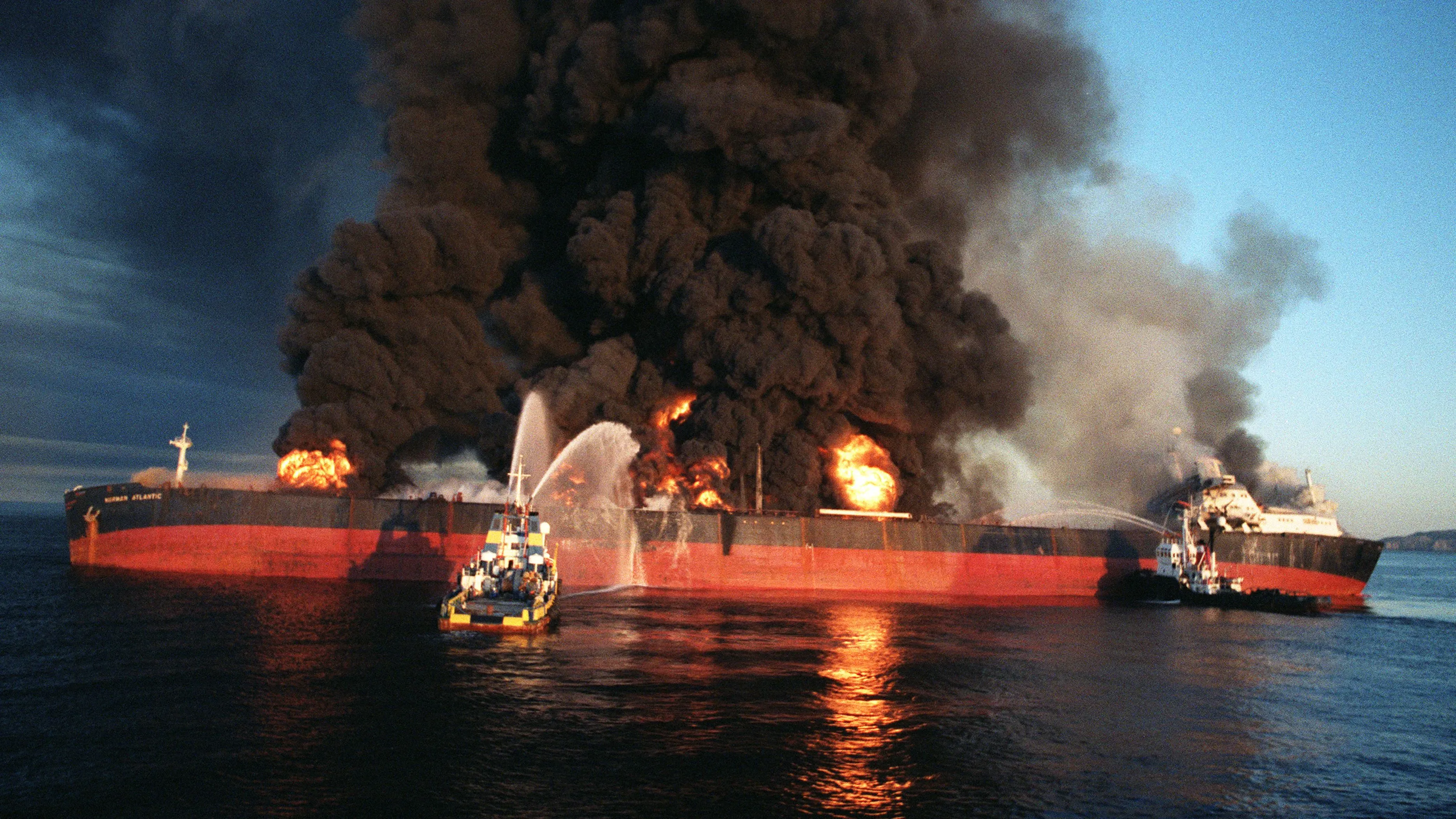 ناقلة نفط مشتعلة في البحر، تتصاعد منها سحب كثيفة من الدخان الأسود، بينما تقترب منها قوارب إطفاء.