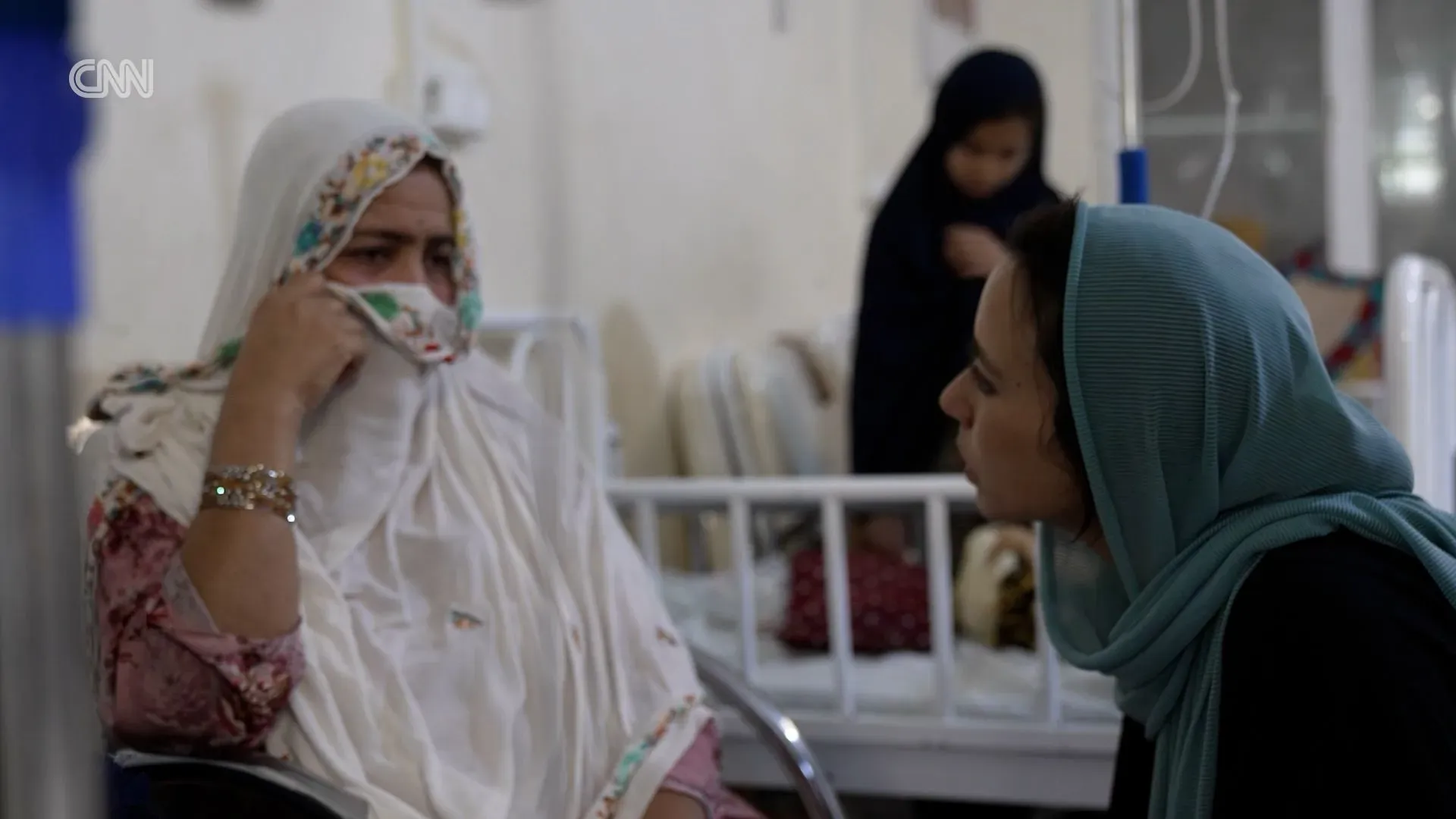 امرأة ترتدي ثوبًا زهريًا تجلس في مستشفى بأفغانستان، تعبر عن ألمها بينما تتحدث مع صحفية، في ظل ظروف صعبة بسبب نقص المساعدات الإنسانية.