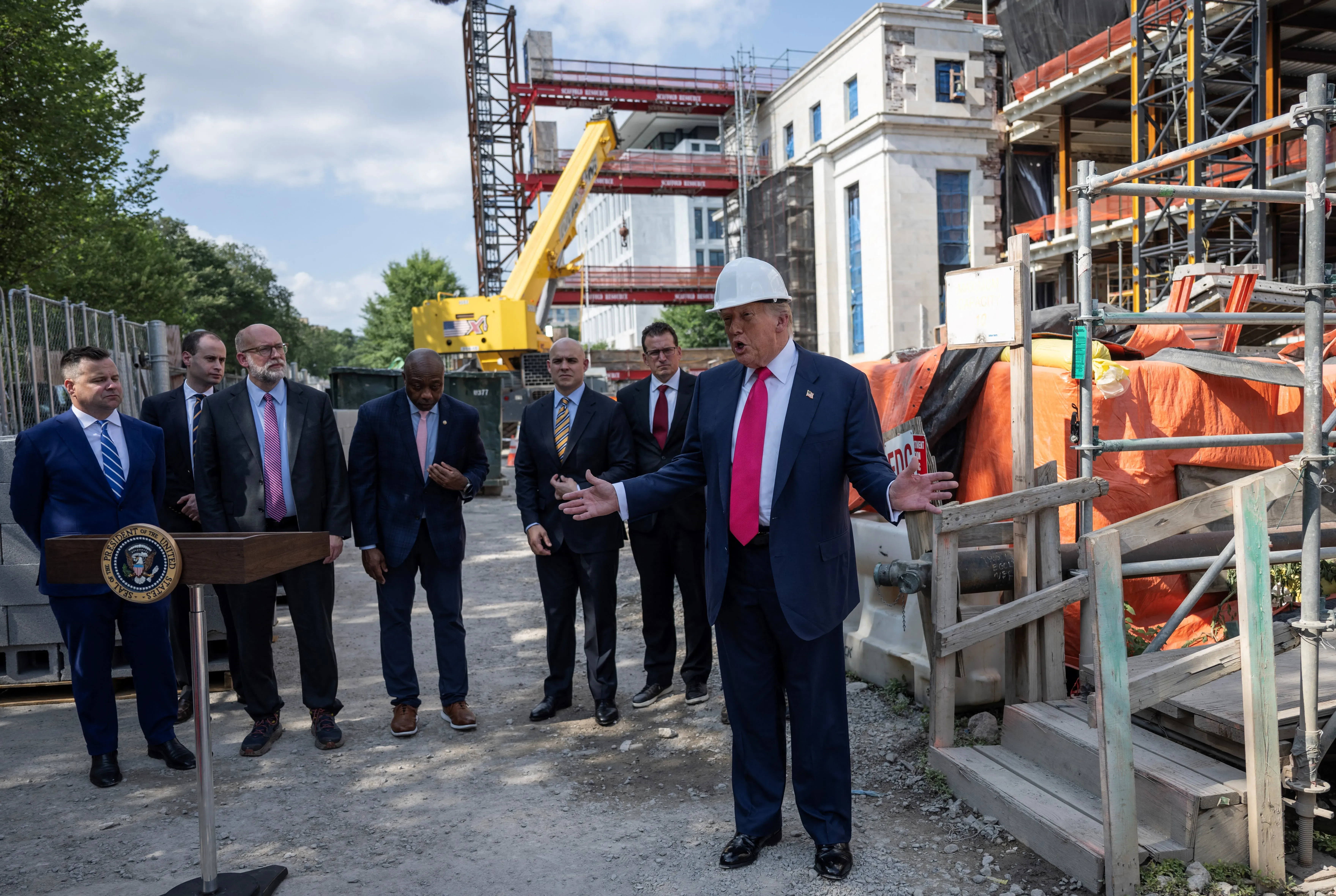 دونالد ترامب يرتدي خوذة بناء ويتحدث خلال جولة في تجديدات مجلس الاحتياطي الفيدرالي، محاطًا بمجموعة من المستشارين.