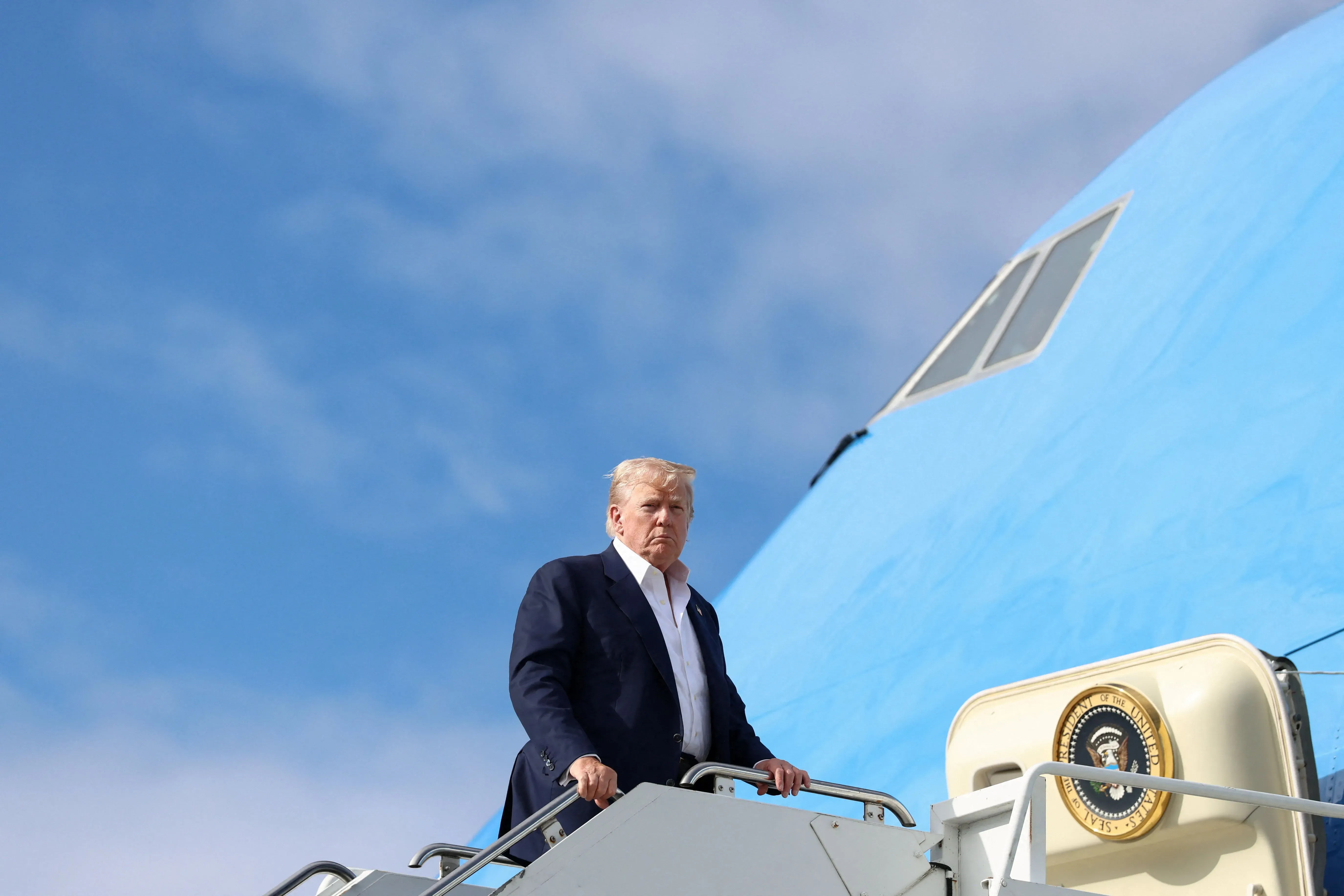 دونالد ترامب يصعد إلى الطائرة الرئاسية، مع خلفية سماء زرقاء، في سياق زيارته لمجلس الاحتياطي الفيدرالي ولقائه مع جيروم باول.