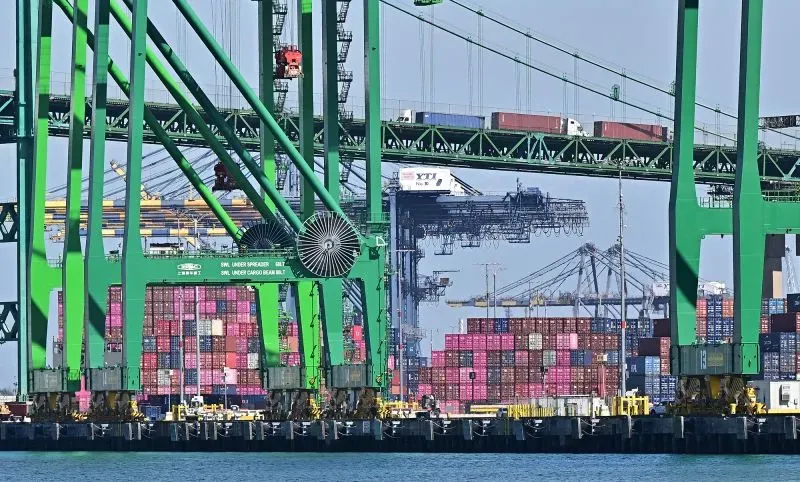 صورة لميناء يحتوي على رافعات خضراء وشحنات ملونة، تعكس النشاط التجاري وتأثير الرسوم الجمركية على الاقتصاد الأمريكي.