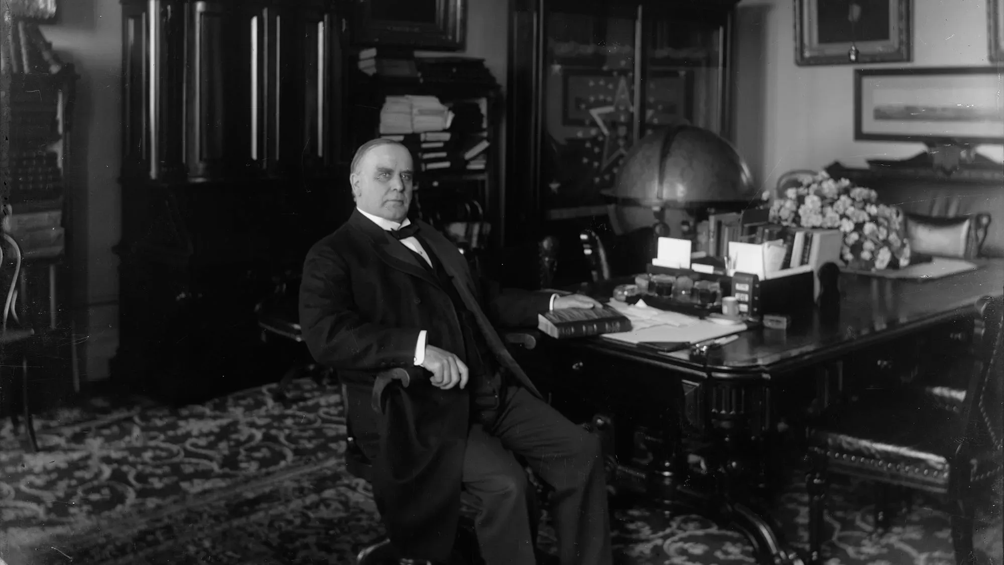 ويليام ماكينلي يجلس في مكتبه، محاطًا بكتب وأدوات مكتبية، مع خريطة العالم خلفه، في سياق حديث عن التعريفات الجمركية.