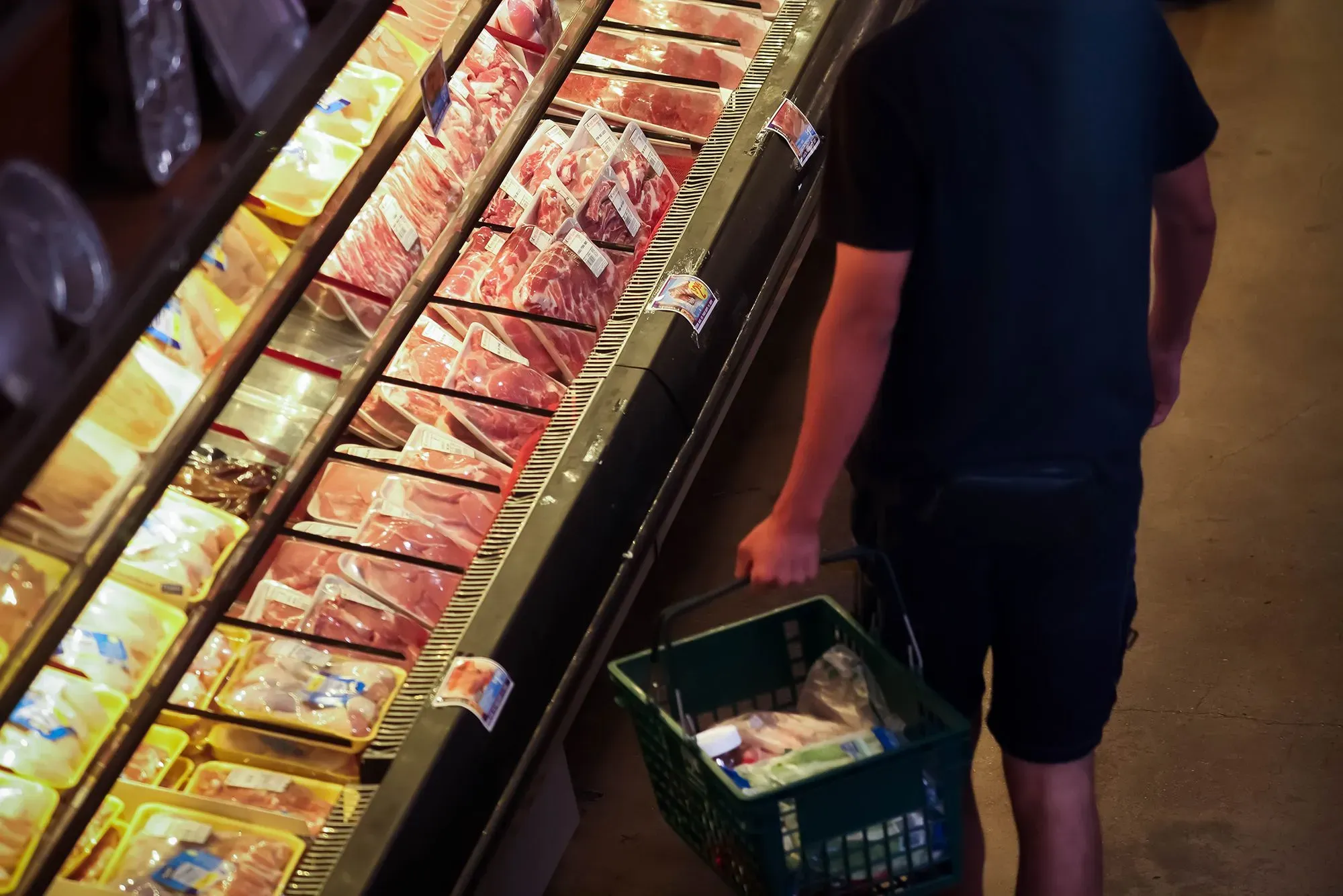 شخص يتسوق في متجر، يحمل سلة ويستعرض مجموعة من اللحوم المعبأة، مما يعكس تأثير التضخم على أسعار المستهلك.