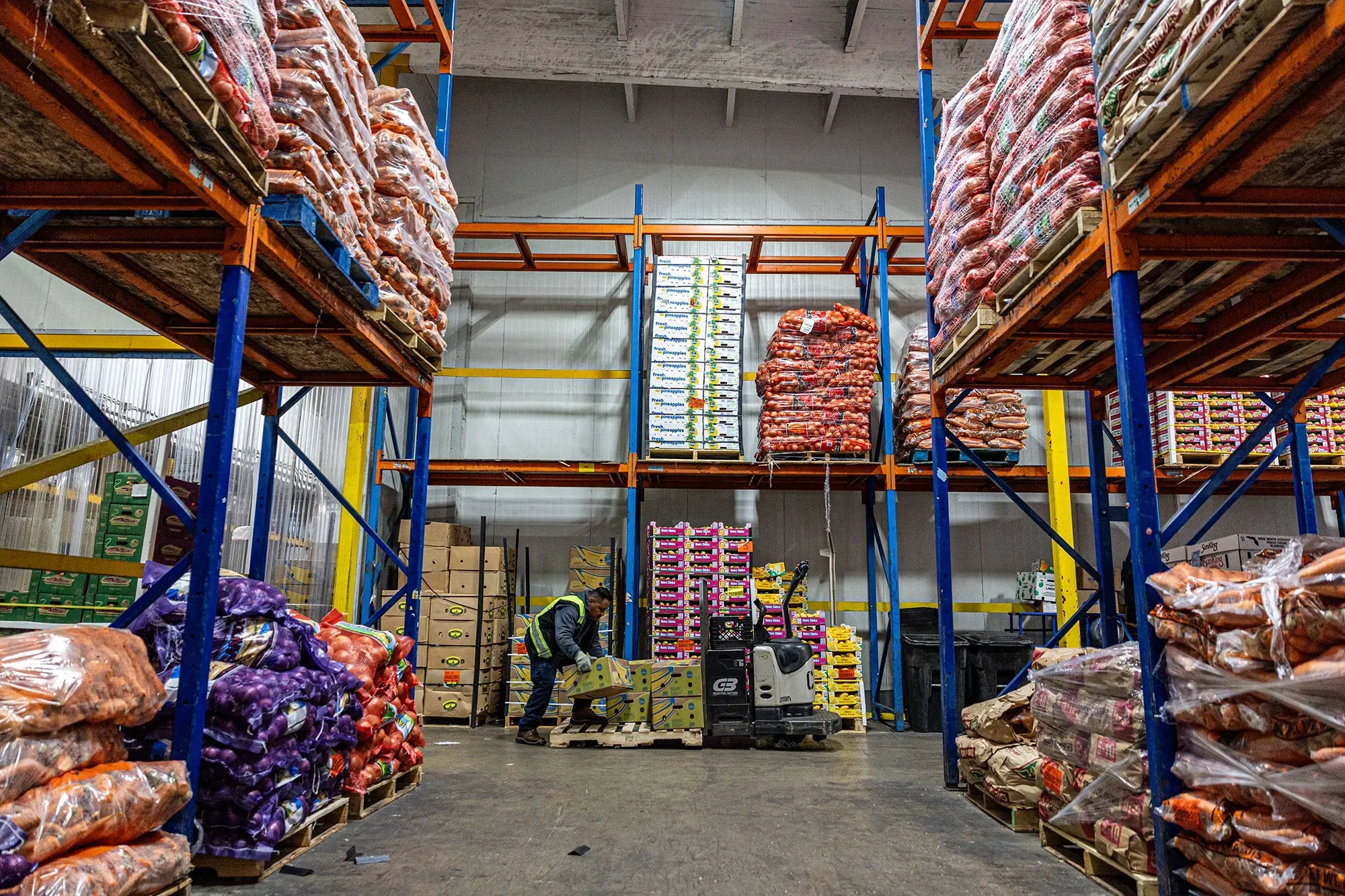 مستودع يحتوي على أكوام من الخضروات والفواكه، مع عامل يقوم بترتيب البضائع، مما يعكس نشاط السوق واستجابة الاقتصاد الأمريكي.