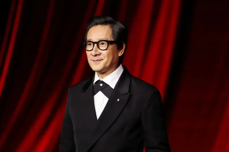 كي هوي كوان يرتدي بدلة أنيقة ويبتسم أمام خلفية حمراء، مما يعكس أجواء الفخامة المرتبطة بمسلسل "اللوتس الأبيض".