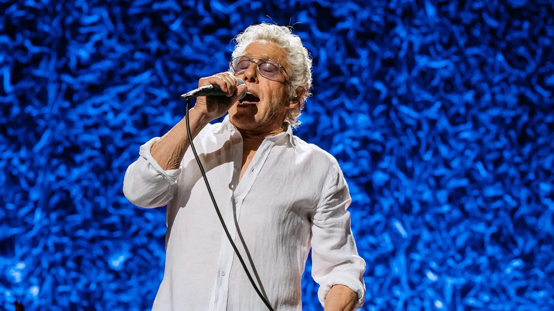 روجير دالتري، مغني فرقة The Who، يؤدي على المسرح في قاعة ألبرت هول الملكية، مع خلفية زرقاء، معبراً عن تحديات التقدم في العمر.