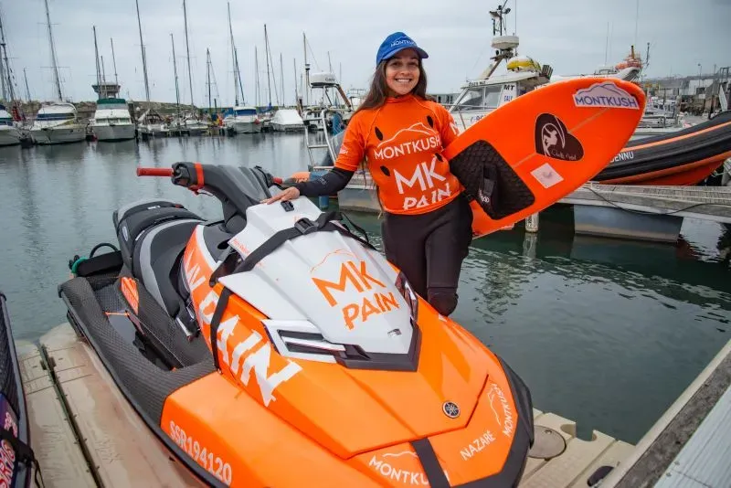 راكبة الأمواج لورا كرين مبتسمة في ميناء نازاري، ترتدي زيًا برتقاليًا وتحمل لوح تزلج، بجانب دراجة مائية، تعكس شغفها في رياضة ركوب الأمواج.