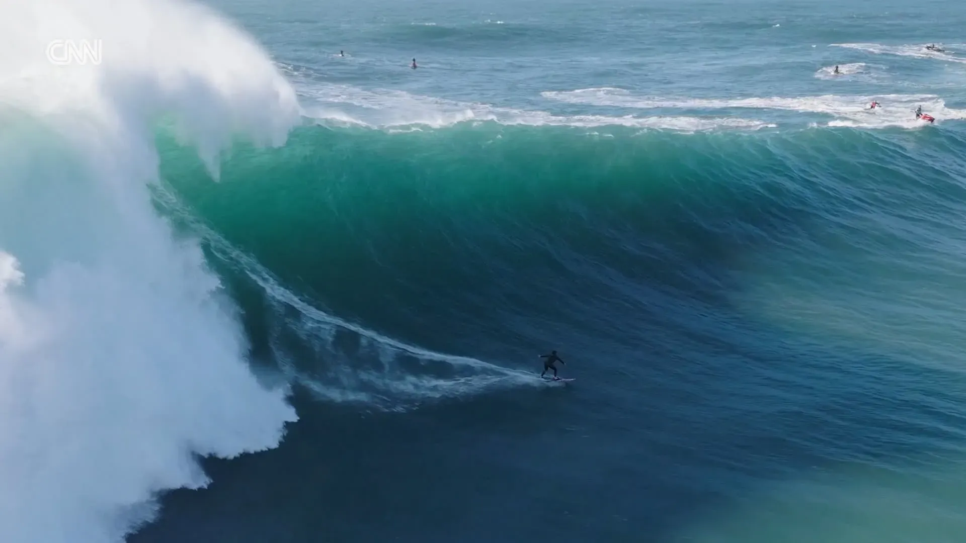 راكبة أمواج تتزلج على موجة عملاقة ارتفاعها 60 قدماً، بينما يظهر خلفها جدار ماء هائل. الصورة تعكس شجاعة وتحدي رياضة ركوب الأمواج.