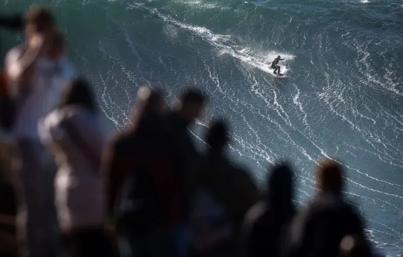 راكبة أمواج تتزلج على موجة عملاقة في نازاري، مع حشد من المتفرجين في الخلفية، تعكس شجاعة النساء في رياضة ركوب الأمواج.