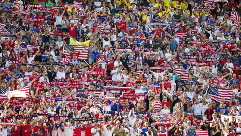 مدرج مليء بالمشجعين يرتدون قمصانًا بألوان مختلفة، يحملون لافتات وأعلامًا، يعبرون عن حماسهم قبل بدء مباراة كرة القدم.