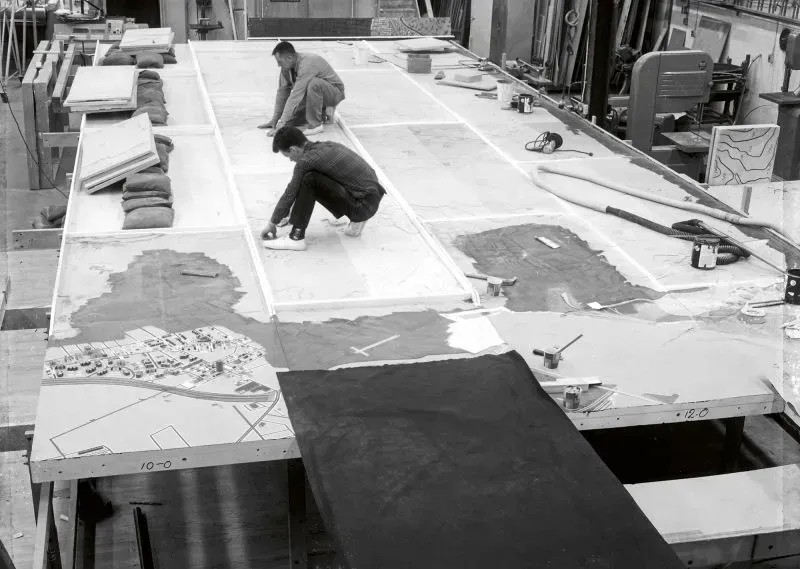 عمال يقومون بتصميم نموذج بانورامي لمدينة نيويورك، يظهرون وهم يضعون تفاصيل دقيقة على السطح الخشبي، مما يعكس الجهد المبذول في المشروع.