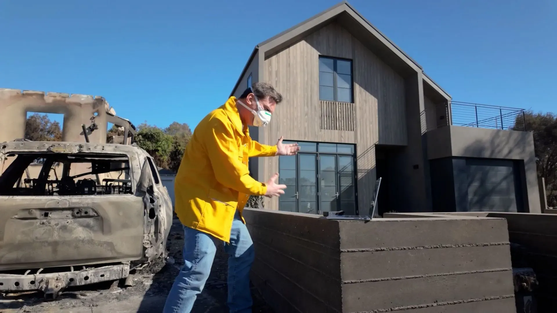 رجل يرتدي سترة صفراء وقناع وجه، يشير إلى الأضرار بسبب حرائق في لوس أنجلوس، مع وجود سيارة محترقة ومنزل جديد بجانبه.