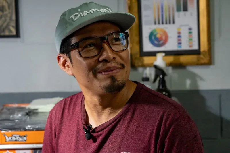 رجل يرتدي قبعة ونظارات، مبتسم في استوديو تاتو، مع أدوات رسم وألوان في الخلفية، يعكس ثقافة الوشوم في السلفادور.