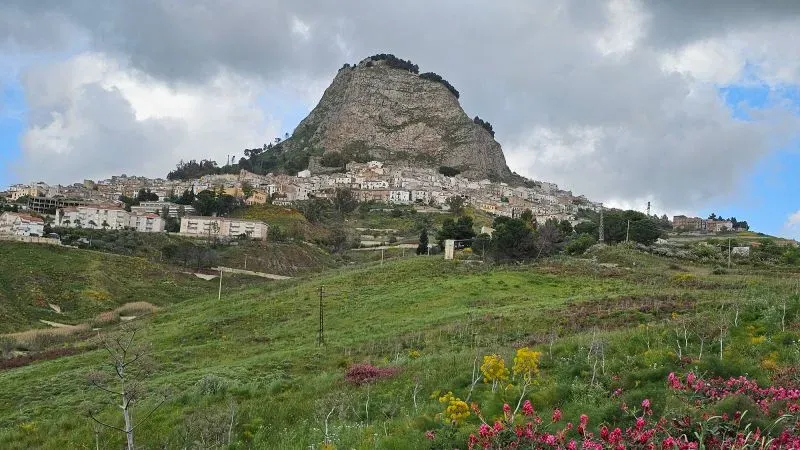 منظر لقرية صقلية القديمة محاطة بالتلال الخضراء، مع جبل بارز في الخلفية، يعكس جمال المنطقة التاريخي وجاذبيتها للمستثمرين في العقارات.