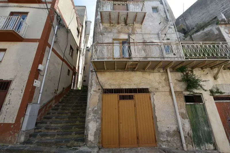 منزل قديم متهالك في بلدة صقلية، يظهر تفاصيل العمارة التقليدية مع شرفة خشبية وألوان باهتة، يعكس فرصة شراء عقار بسعر يورو واحد.