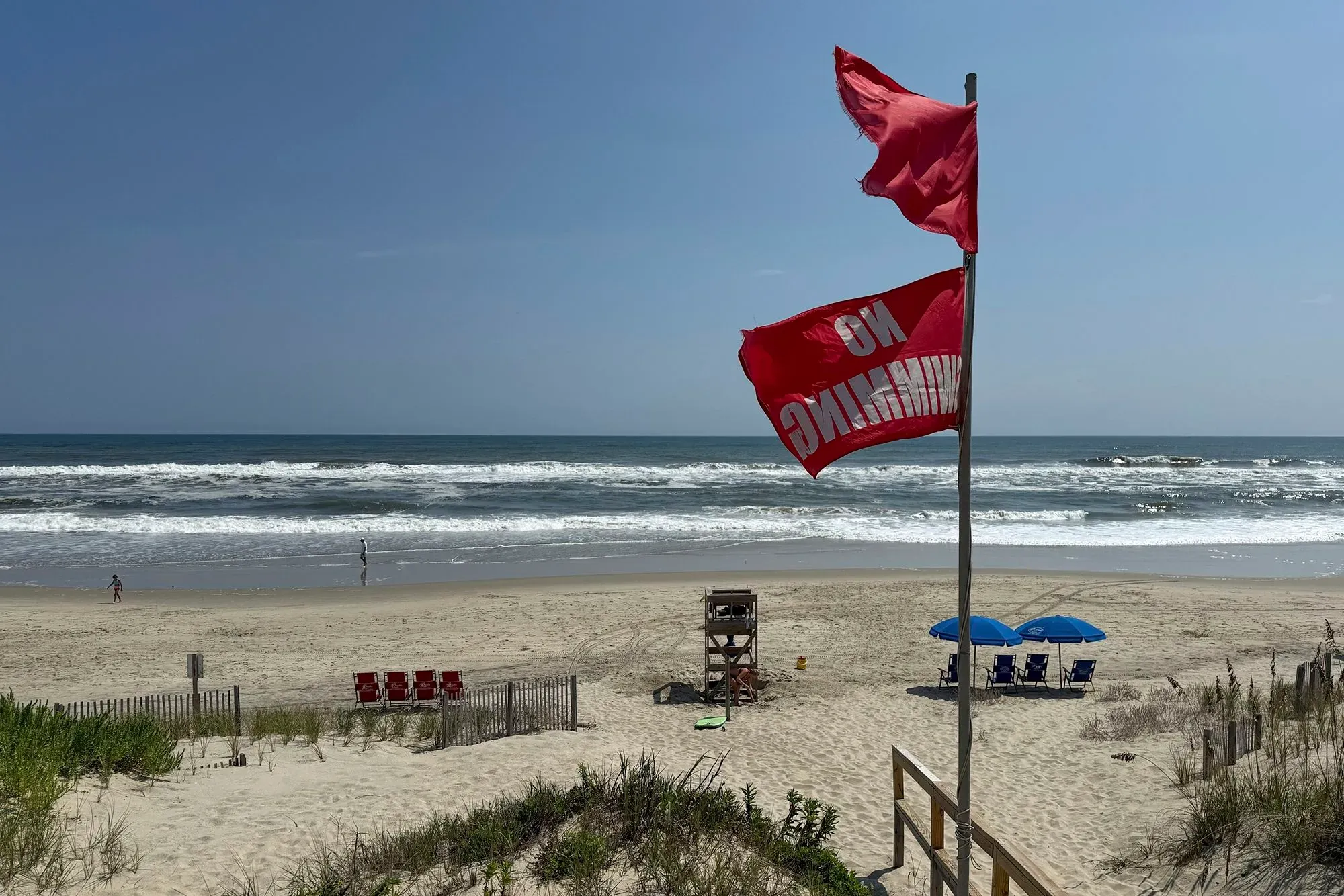 علم أحمر مكتوب عليه "لا سباحة" يرفرف فوق شاطئ هادئ، حيث تظهر الأمواج العالية والرمال، مما يعكس خطر الإعصار إيرين.