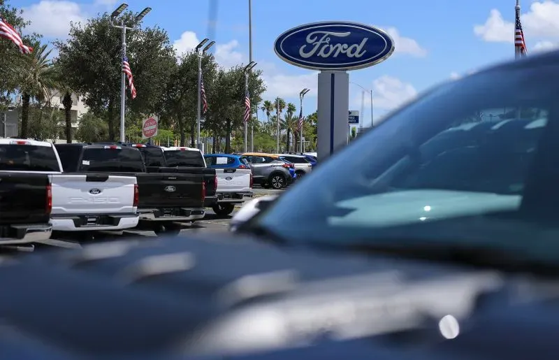 شعار شركة فورد يظهر في مركز بيع سيارات مع صف من السيارات في المقدمة، مما يعكس تأثير التعريفات الجمركية على أسعار السيارات.