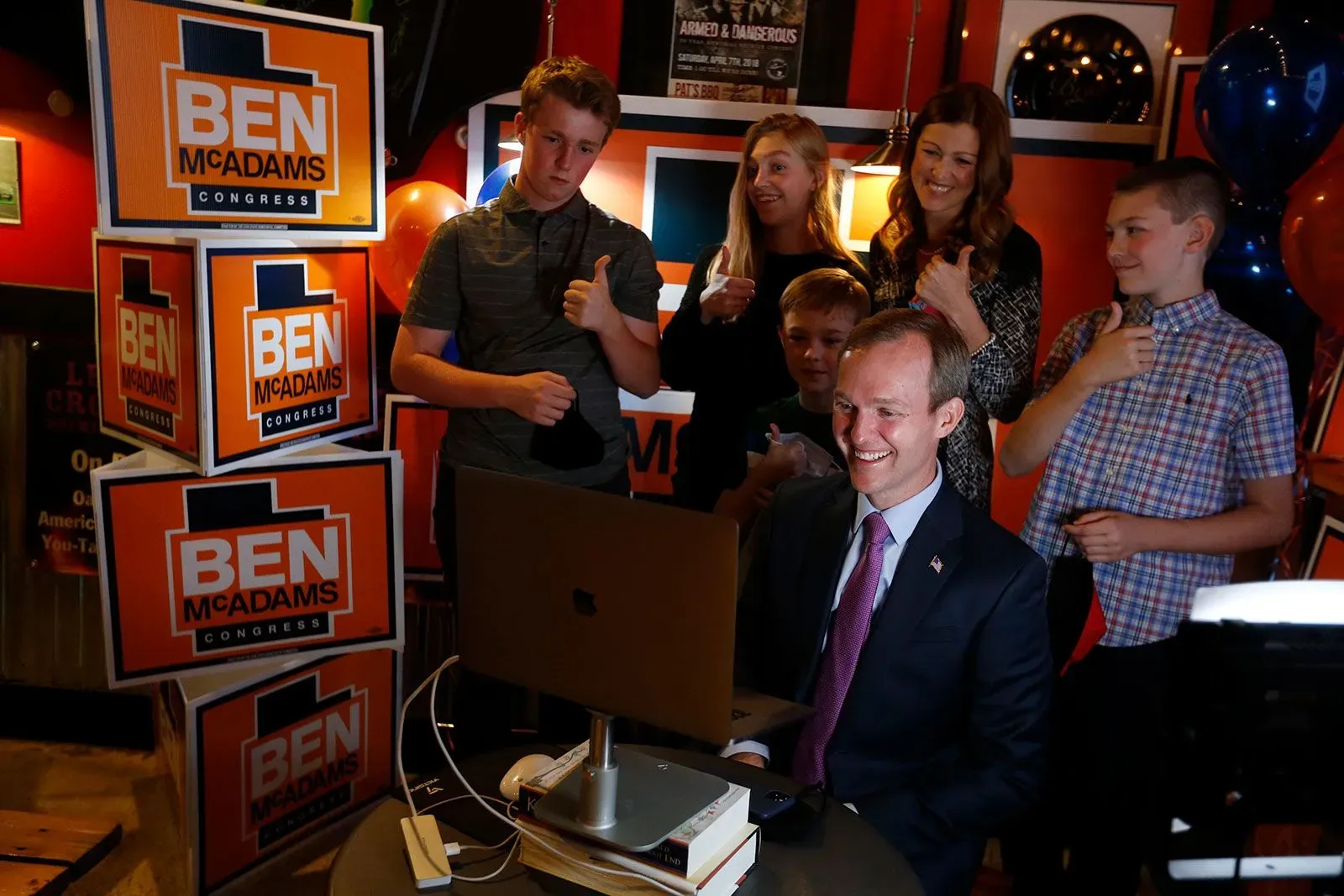 بن ماك آدامز مبتسم أمام جهاز كمبيوتر محمول خلال حدث انتخابي، محاطًا بأفراد عائلته وأصدقائه الذين يظهرون دعمهم له.