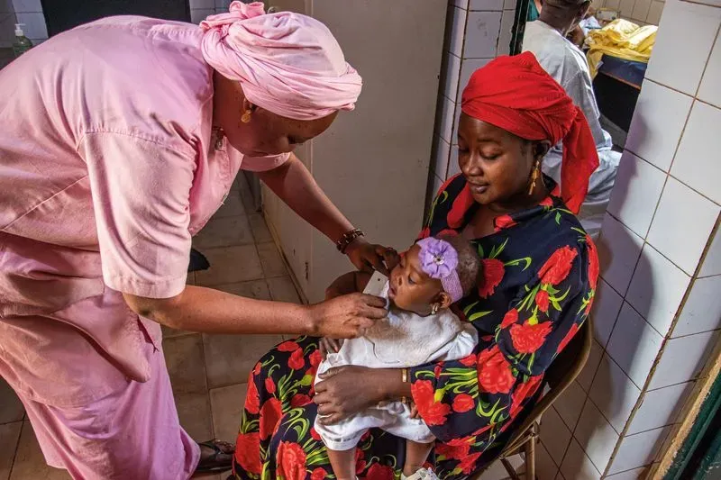 امرأة ترتدي ملابس تقليدية تحمل طفلاً بينما تقوم مربية بفحص صحة الطفل في مركز طبي، في سياق جهود مكافحة سوء التغذية في نيجيريا.