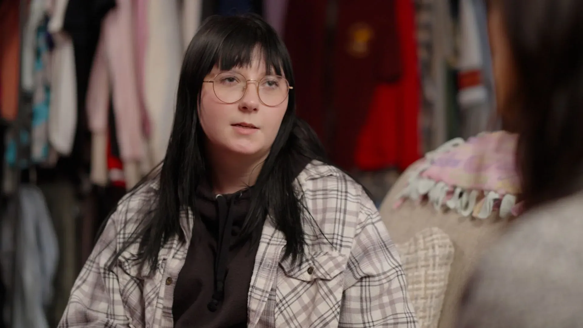 فتاة شابة ترتدي سترة مربعة وتجلس في غرفة مليئة بالملابس، تعبر عن مشاعرها حول موضوع حسّاس يتعلق بالجرائم المدرسية.