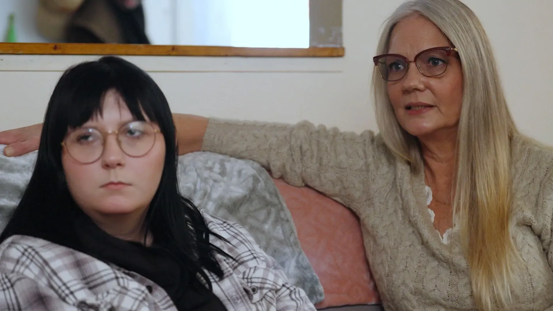 امرأة ذات شعر طويل ومجعد ترتدي نظارات، تجلس بجانب فتاة شابة ذات شعر أسود قصير، ترتدي نظارات أيضاً. تعبر ملامح وجههما عن التأمل والقلق.