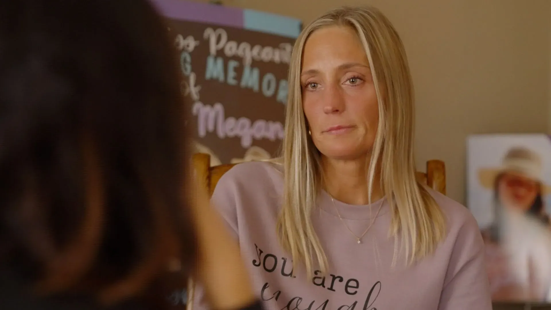 امرأة شابة تجلس في غرفة مع خلفية تحتوي على عبارات ملهمة، تعبر عن مشاعر الحزن والقلق بعد فقدان ابنتها.