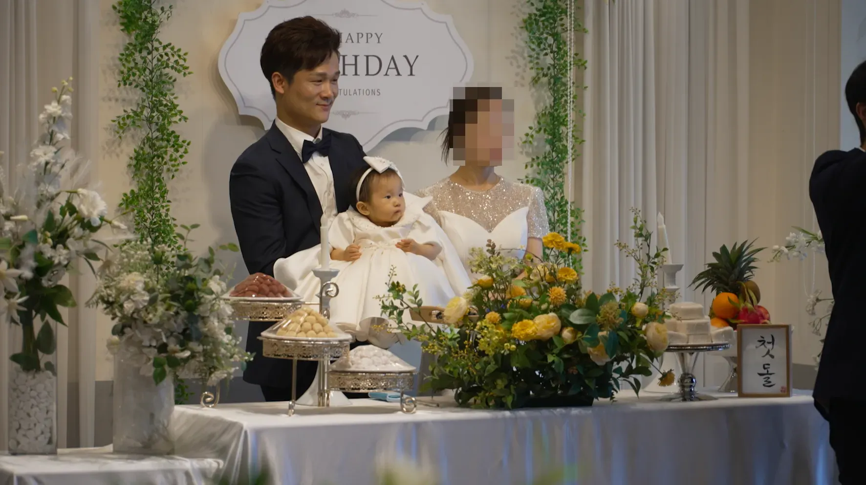 احتفال بعيد ميلاد طفل يرتدي ملابس تقليدية، مع والدين في ملابس رسمية، وزينة جميلة تشمل الزهور والحلويات، تعكس تقاليد الفرح والاحتفال.