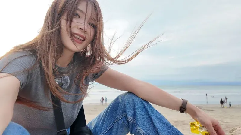 امرأة شابة تجلس على الشاطئ، مبتسمة وتستمتع باللحظة. يظهر في الخلفية مسافرون آخرون وسماء زرقاء. تعكس الصورة تجربة السفر والمغامرة.