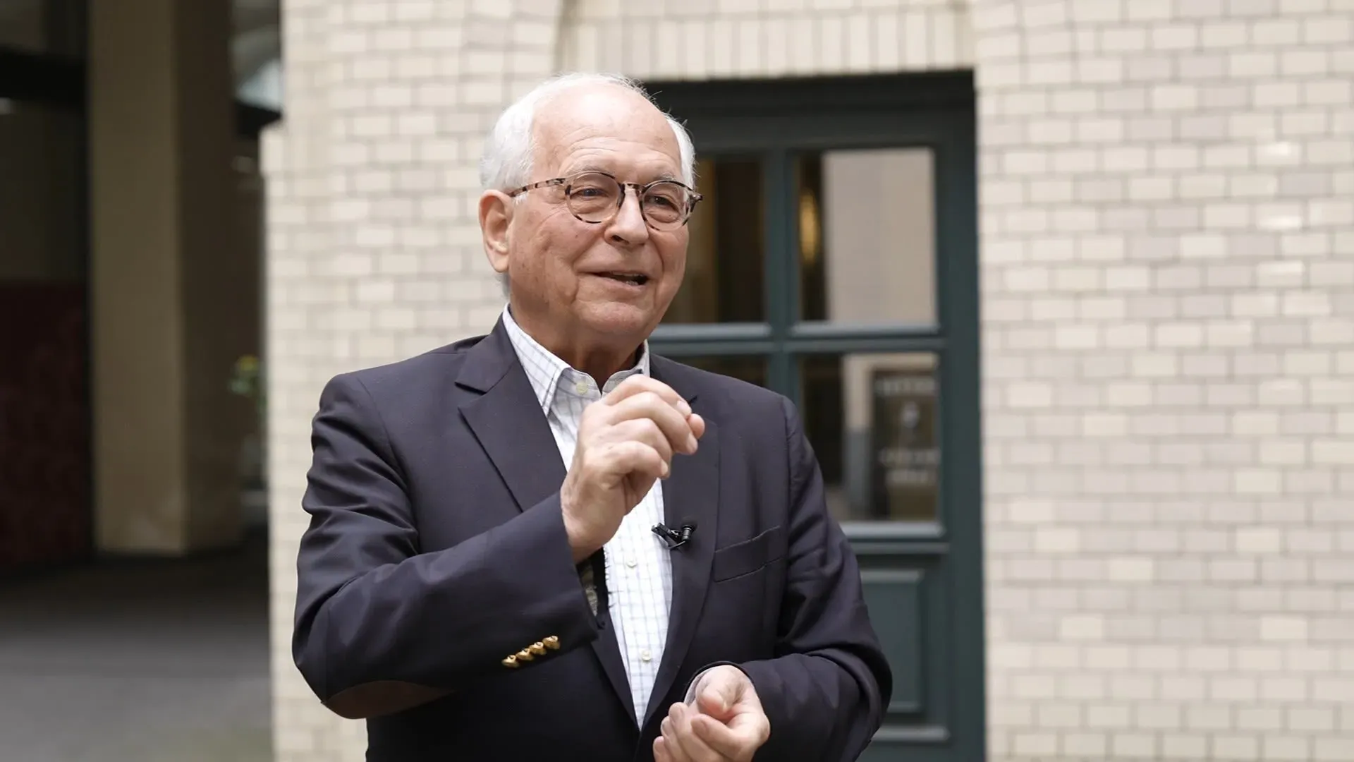 رجل مسن يرتدي نظارات وقميصًا أزرقًا، يتحدث بحماس أمام مبنى ذو جدران من الطوب الأبيض، مع نافذة خلفه، مما يعكس روح التعاون الأوروبي بين فرنسا وألمانيا.