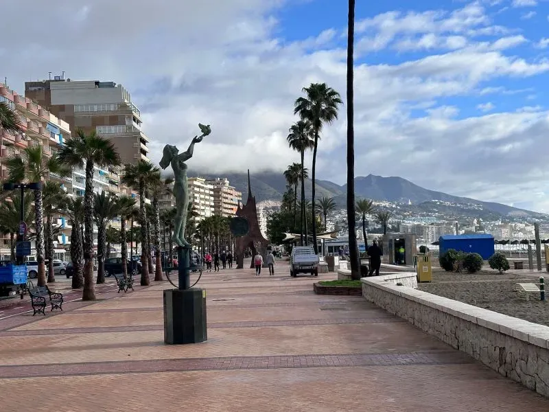 شارع في مدينة فوينخيرولا بإسبانيا، يضم تمثالاً و Palm trees، مع منظر للجبال في الخلفية، يعكس الحياة الهادئة على ساحل البحر الأبيض المتوسط.