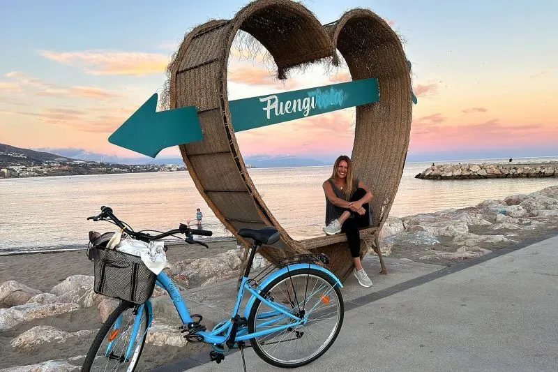 امرأة تجلس داخل إطار قلب كبير مكتوب عليه "Fuengirola" بجوار دراجة زرقاء، مع منظر للبحر وغروب الشمس في الخلفية.