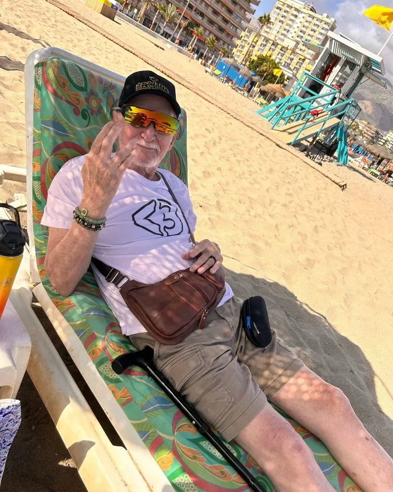 رجل مسن يجلس على شاطئ رملي في فوينخيرولا، يرتدي نظارات شمسية وقبعة، ويشير بإشارة اليد، مع خلفية تضم مباني وشجر النخيل.