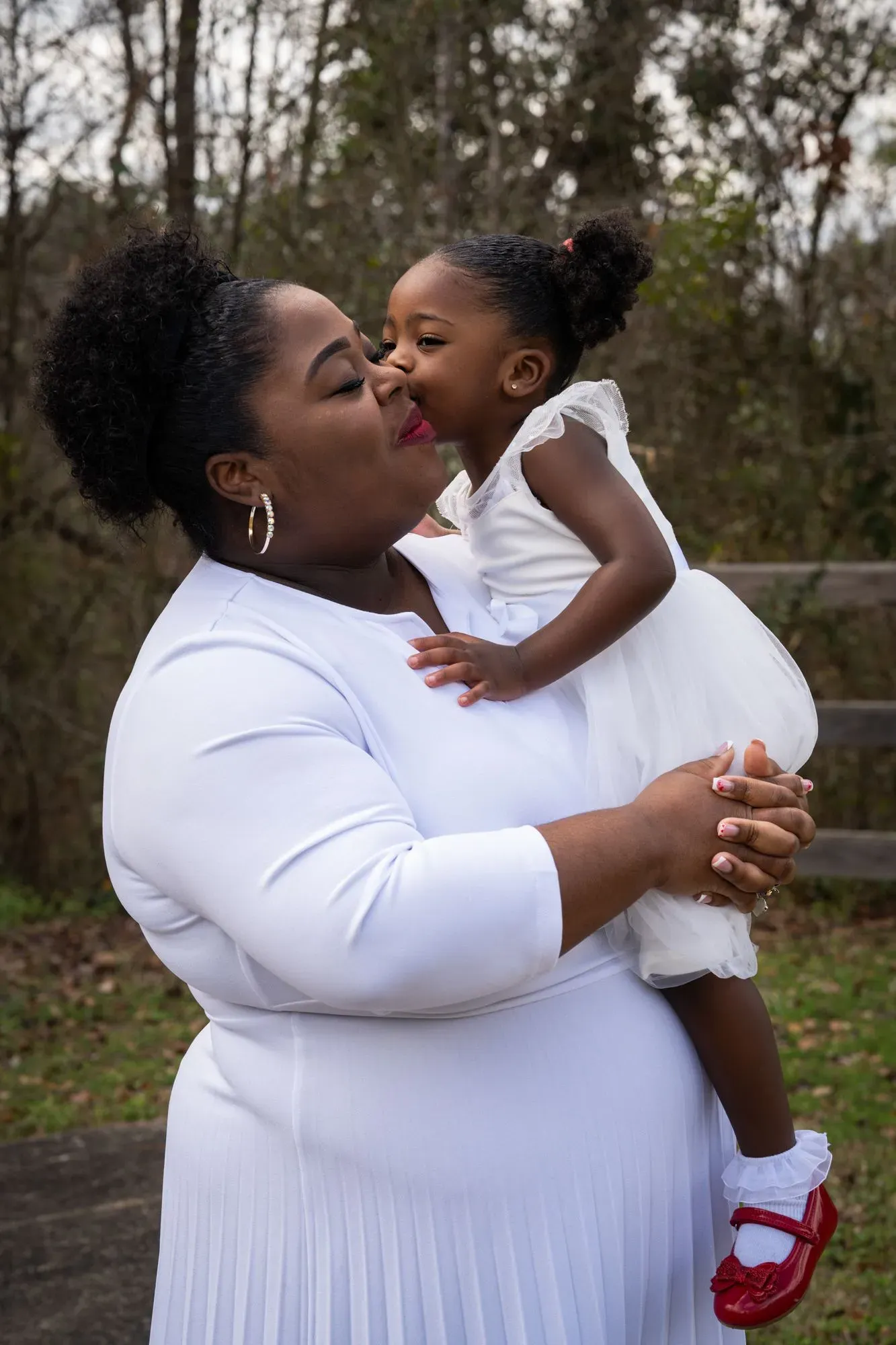 امرأة حامل ترتدي فستانًا أبيض، تحمل طفلة صغيرة ترتدي فستانًا أبيض أيضًا، يتبادلان القبلات في بيئة طبيعية. تعكس الصورة روابط الأمومة والعناية.