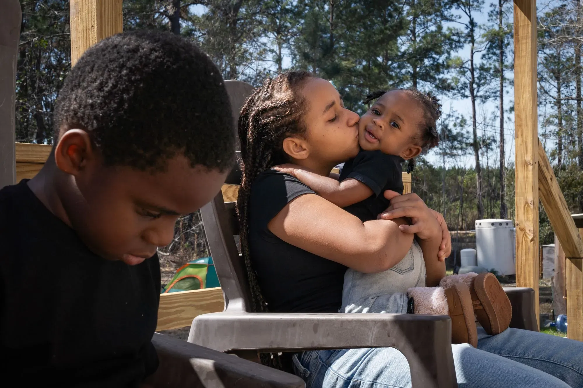 امرأة تحتضن طفلًا وتقبله، بينما يجلس طفل آخر بجانبها. تعكس الصورة لحظة عائلية دافئة في بيئة طبيعية.