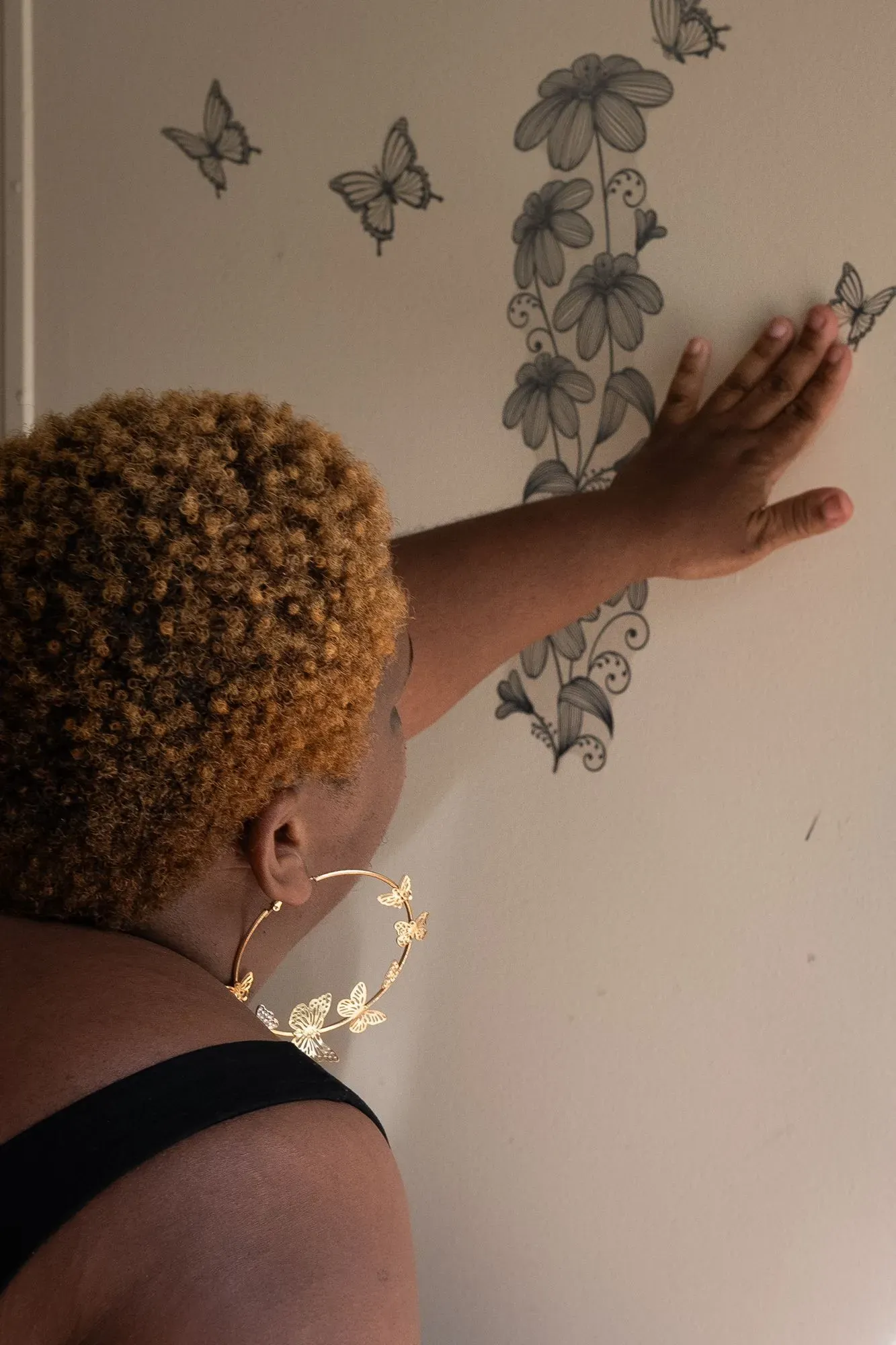 امرأة ذات شعر قصير ومجعد تلمس جدارًا مزينًا برسومات زهور وفراشات، تعبر عن لحظة من التأمل أو التفكير.